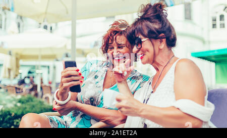 Zwei glückliche älterer Frauen Freunde über ein Smart Phone und sprechen beim Sitzen draußen in einem Café Stockfoto