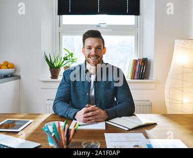 Schöner Lächeln bärtiger Mann mit gefalteten Händen in legere Kleidung am Schreibtisch sitzen vor dem hellen Fenster Stockfoto