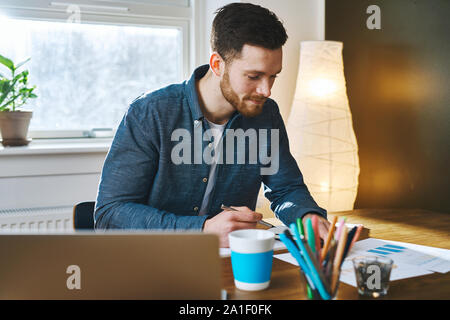 Eine ernsthafte junge Bartgeier erwachsenen männlichen Arbeiten am Schreibtisch im Büro zu Hause mit weichen Stehleuchte hinter ihm am Fenster Stockfoto