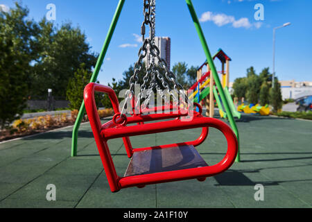 Leeres rotes schaukeln auf dem Spielplatz gegen den blauen Himmel und Bäumen.. Swing ist hängen an den Ketten aus Metall. Ein Ort für Kinder, um Spaß zu haben. Sonnigen Tag. Stockfoto