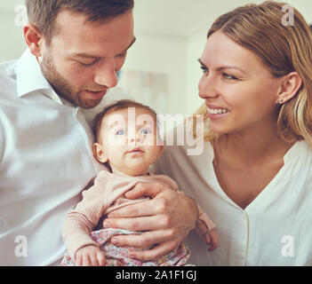 Glückliche junge Mutter und Vater zusammen an einem Tisch zu Hause sitzen schaukelnd ihre neugeborene Tochter Stockfoto