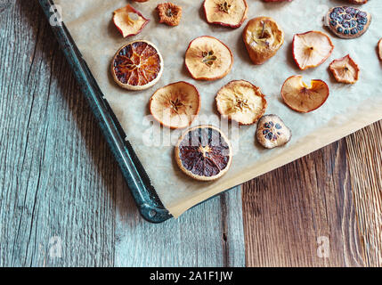 Hausgemachte Scheiben getrocknete Orangen und Äpfel auf Backpapier und schwarz Backblech, diagonale Anordnung von oben rechts, Holz- Hintergrund, Angl Stockfoto