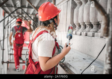 Weibliche bau Maurer steht hoch auf der Gerüste, Arbeiten mit Silikon Pistole und der Renovierung der Wand auf der alten Versorgungsgebäude Stockfoto