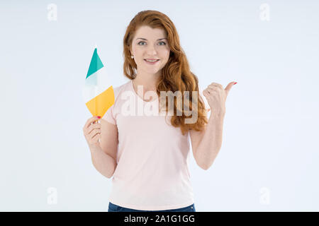 Rothaarige junge Frau mit der Flagge von Irland weist mit dem Finger auf die Seite und stand auf einem hellen Hintergrund. Stockfoto