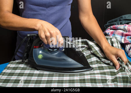 Frau in einem blauen T-Shirt bügeln Plaid Shirt und andere Bekleidung mit schwarzem Eisen auf einem dunklen Hintergrund Stockfoto