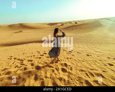 Junge schöne Frau in Kleid und mit einem Hut in der Mitte des Dubai Wüste mit Sonnenlicht. Stockfoto