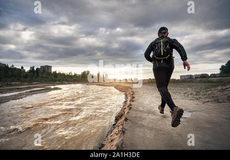 Mann in Schwarz Shirt mit Rucksack, der auf der dreckigen Trail in der Nähe des Flusses bei Sonnenuntergang Himmel. Gesunder Lebensstil Konzept Stockfoto