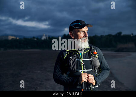 Portrait von älteren Athleten Mann mit grauem Bart und Stöcke an dunklen bewölkter Himmel Hintergrund Stockfoto