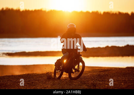 Der Motorradfahrer fährt auf elektrische enduro Motorrad gegen den Fluss und den Sonnenuntergang Licht Stockfoto