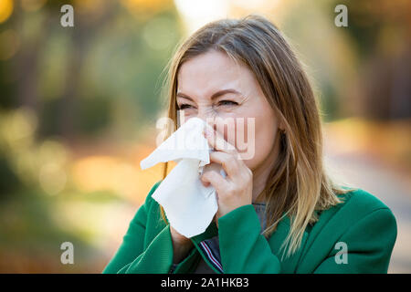 Kranke junge Frau mit Kälte und Grippe stehen im Freien, Niesen, wischen Nase mit Taschentuch, Husten. Herbst Straße Hintergrund Stockfoto