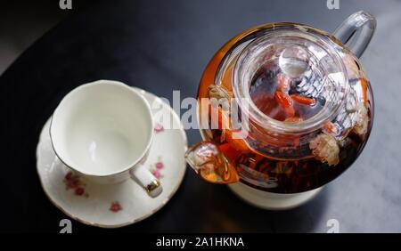 Schwarzer Tee mit goji (chinese wolfberries) und Jasmin Teekanne aus Glas und Porzellan Tasse mit Untertasse Blumenmuster auf schwarz Tabelle Stockfoto