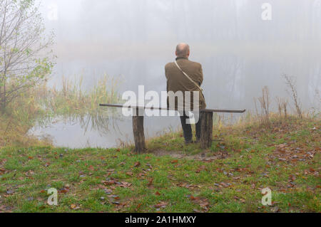 Älterer Mann sitzt einsam auf einer Bank am Flußufer auf Misty herbstliche monring Stockfoto