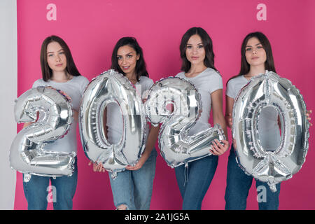 Eine Gruppe von schönen Mädchen in weißen T-Shirts und Jeans sind holding Air silber Kugeln in der Form von Zahlen 2020 auf einem hellen rosa Hintergrund Stockfoto