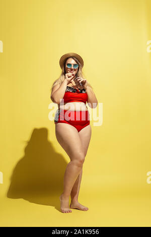 Junge kaukasier plus Größe weibliche model in Vorbereitung auf die Beach Resort auf gelben Hintergrund. Frau in rot Badeanzug, Hut und Sonnenbrille. Konzept der Sommer, Party, Körper positiv. Cocktail trinken. Stockfoto