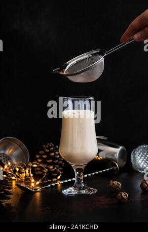 Eierlikör traditionelle Weihnachten alkoholisches Getränk mit Zimt und Muskatnuss. Hand bestreut Zimtpulver auf Glas. Winterurlaub Stimmung Stockfoto