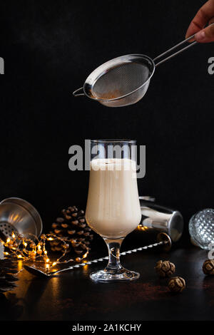 Eierlikör traditionelle Weihnachten alkoholisches Getränk mit Zimt und Muskatnuss. Hand bestreut Zimtpulver auf Glas. Winterurlaub Stimmung Stockfoto