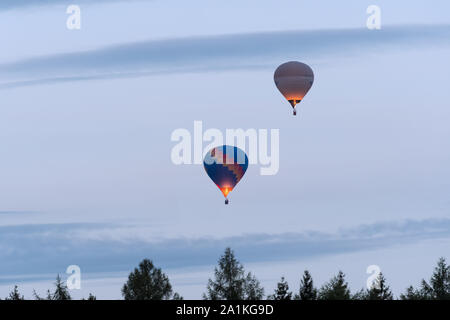 Zwei Heißluftballons schweben in den bewölkten Himmel. Im Vordergrund, Treetops sind sichtbar, als Silhouetten. Gas Antrieb Feuer flammen. Stockfoto