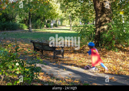 Ein kleines Mädchen auf einem Roller flitzt durch, in einem Super Held Kostüm an Stonegrove Park im Herbst gekleidet. Stockfoto