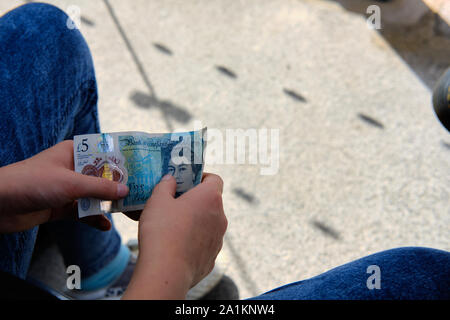 Leeds/UK - Juli 22 2019: Ein junger Mensch sitzt mit fünf Pfund Sterling Kunststoff Hinweis in Ihrer Hand an einem sonnigen Tag. Stockfoto