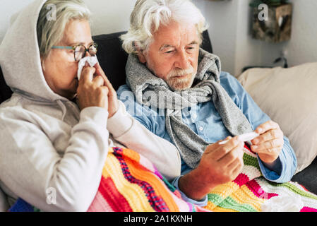 Paar sick Senioren und ältere Menschen auf dem Sofa sitzen mit Fieber auf das Thermometer mit hoher Temperatur suchen - zwei ältere Frau und Mann Stockfoto