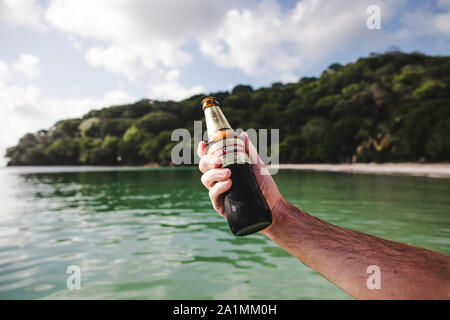 Kaukasische männliche Hand, die eine erfrischend kalte Flasche des Club Colombia - das erstklassige nationale Bier Kolumbiens - an einem Strand auf einer karibischen Insel hält Stockfoto