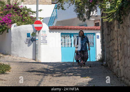 Alacati, Türkei - 4. September 2019: Alte Frau mit Stöcken wandern in einer engen Straße. Die Stadt ist bei Touristen beliebt im Sommer Stockfoto