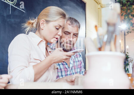 Glücklich verheiratetes Paar am Tisch sitzen in der Töpferei. Frau macht Foto auf dem Smartphone. Stockfoto