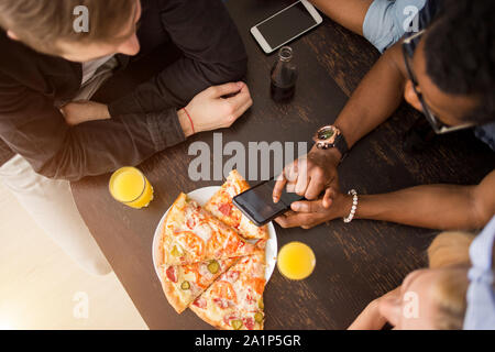 Blick von oben auf die Gruppe der multirassischen Freunde sitzen um den Tisch, Mittagessen, essen Pizza, Saft trinken, über Handy. Lieferung, Essen, Technol Stockfoto