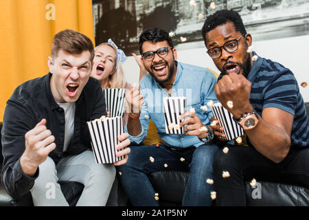 Fröhlich aufgeregt Gruppe der multirassischen Freunde sitzen auf der Couch essen Popcorn, watching Sport Spiel im Fernsehen, schreien, feiern Ziel. Freizeit, Sport, Stockfoto