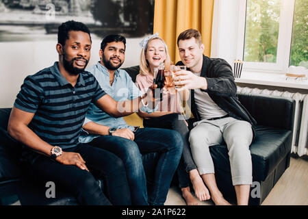 Fröhlich multikulturellen Freunde Studenten legere Kleidung trinken, klirrenden Flaschen und Gläser Getränke, feiert Geburtstag. Drei Mann und woma Stockfoto