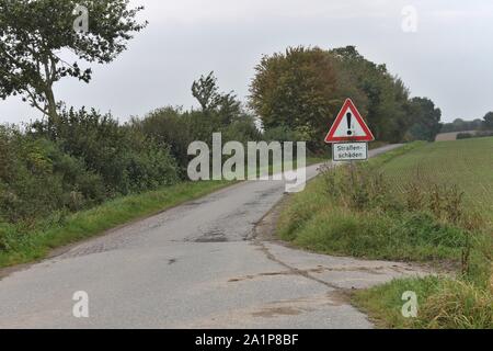 Schild Warnung der Fahrbahnoberfläche Schaden in deutscher Sprache. Am Straßenrand Schäden sichtbar sind, beginnend Neben dem Schild. Stockfoto