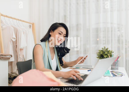 Fashion Designer bei der Arbeit. Glückliche junge asiatische Frau Zeichnung beim Sitzen an Ihrem Arbeitsplatz in Fashion workshop Stockfoto