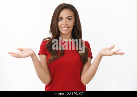 Warum so ernst. Überrascht junge süße Brünette Frau mit Sommersprossen im roten T-Shirt kann nicht verstehen, was geschieht, mit einem Schulterzucken Hände heben u in Frage gestellt Stockfoto