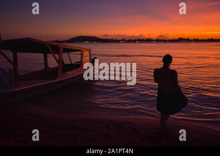 Junge Frau mit Blick auf Gili Trawangan als im Hintergrund, Bali, Indonesien von Gili Meno mit Mount Agung nach Sonnenuntergang gesehen. Stockfoto