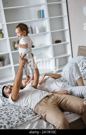 Freundliche Vater tun Übungen mit Kind, sein Kind heben, wjhile liegend auf Bett mit seiner Frau. Nahaufnahme Seite Foto anzeigen Stockfoto