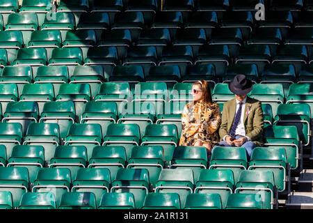 Gut gekleidetes Ehepaar wartet in eine leere Tribüne für Track Aktion im Sonnenlicht am 2019 Goodwood Revival, Sussex, UK. Stockfoto