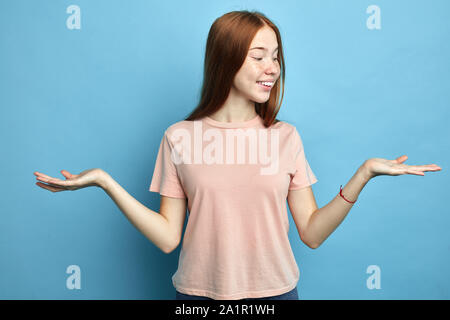 Positiv glückliche Frau wirft beide Handflächen, der vorgibt, die zwei Elemente, die sich in gute Laune, trägt Rosa T-shirt, auf blauen Hintergrund isoliert. S Zufrieden Stockfoto