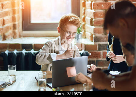 Chherful blonde Junge in Gläsern und sein Team zusammen, die am Projekt, bis schließen Foto Stockfoto