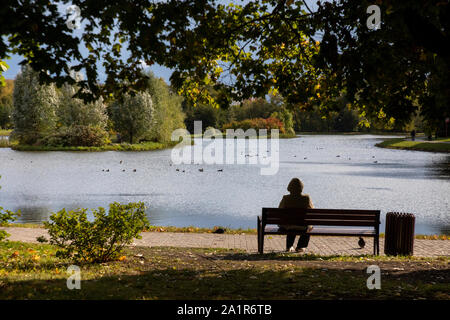 Eine Frau liegt auf einer Bank mit Blick auf einen Teich an einem sonnigen Herbsttag Stockfoto