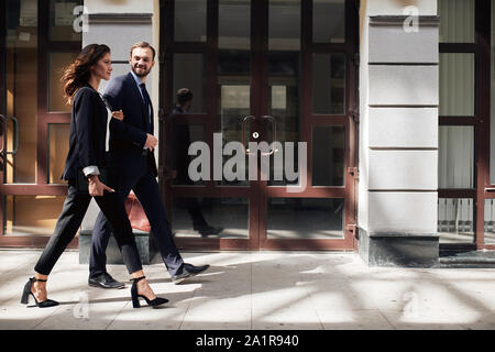 Blonde hübscher Kerl in brunette Schöne Kollegin beim Gehen mit ihr Büro, volle Länge Seite Foto anzeigen Stockfoto