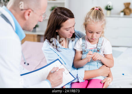 Junge sorgfältige brünette Frau mit ihrer kranken Tochter auf Knien Arzt zusammen Stockfoto