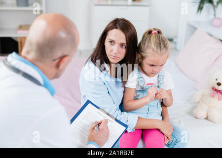 Sorgen junge Mutter hören Hinweise für den Arzt halten sie ihre kranke Tochter auf Knien im Krankenhaus Stockfoto