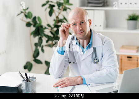 Schwere Arzt in Jungtieren und Brillen zu Ihnen schauen, während Sie von Arbeitsplatz im Krankenhaus sitzen Stockfoto