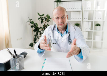 Selbstbewussten jungen Arzt in Jungtieren und Brillen erklären, was Sie beim Sitzen durch Arbeitsplatz Stockfoto