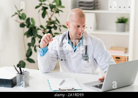 Kahlen Arzt in Uniform durch on-line-Informationen suchen, während sie von Schreibtisch vor Laptop Stockfoto