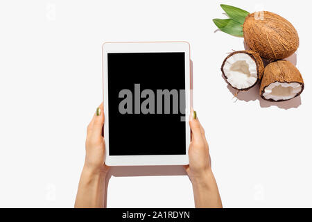 Weibliche Hände halten tablet device mit leeren Bildschirm neben Kokosnüsse und grüne Blätter auf weißem Hintergrund, flach. Stockfoto