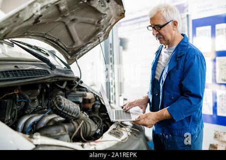 Älterer Mann in der blauen Uniform stehend, die durch den Motor der Maschine beim Suchen im Netz für Anweisungen Stockfoto