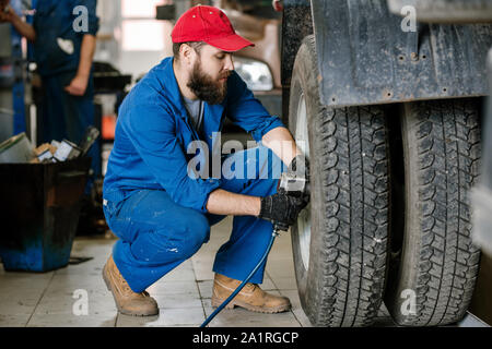 Ernsthafte junge Techniker in Arbeitskleidung sitzen durch das große Rad des Fahrzeugs während Reparaturarbeiten Stockfoto
