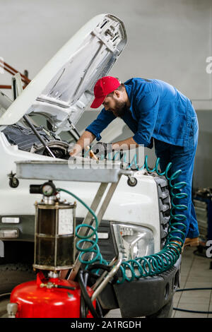 Junge Techniker in Arbeitskleidung bücken Öffnen Motor für große Fahrzeuge, während sie Spiralkabel bei Reparaturarbeiten Stockfoto