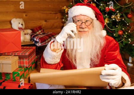 Santa Claus Kostüm, Handschuhe und Brille Holding abgerollt, Papier mit Kindern Namen und im Gespräch mit ihren Eltern auf dem Mobiltelefon Stockfoto